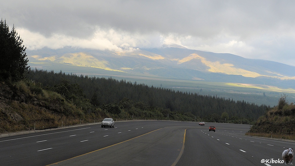 Das Bild zeigt eine Breite sechsspurige Straße mit Standstreifen und beitem Milltelstreifen in einer abschüssigen Rechtskurve. Nur drei wetere Fahrzeuge sind im Bild. Im Hintergrund ist ein hoher Berg, der durch Wolken verdeckt ist.