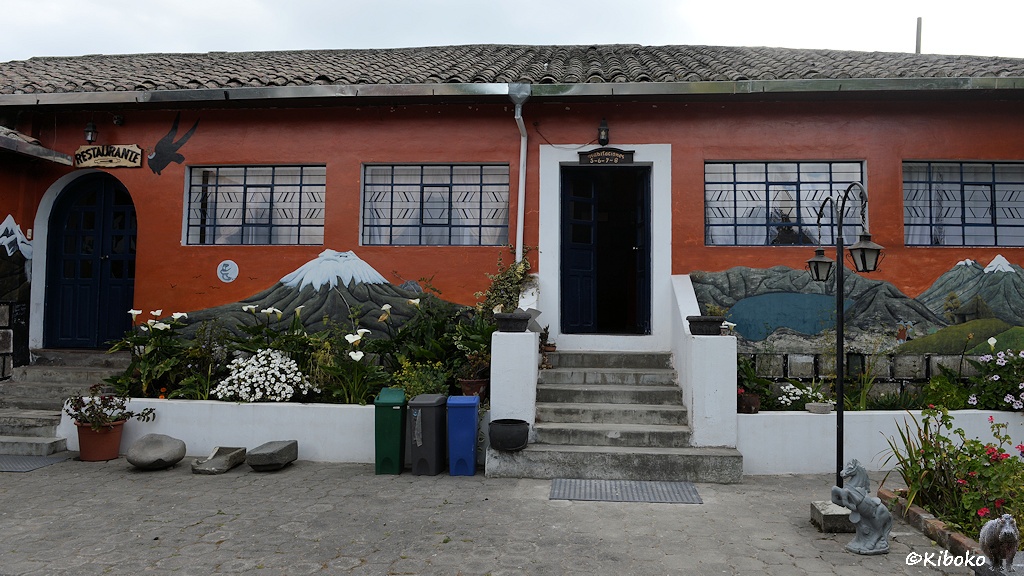 Das Bild zeigt ein rotbraunes Haus mit zwei Türen. Über der linken Tür steht Restaurant. Jeweils sieben Stufen sind vo rden Türen. Davor ist ein kleiner Garten mit weißen und roten Blumen. Am unteren Teil der Hauswand sind Vulkane und Kraterseen der Umgebung gemalt.