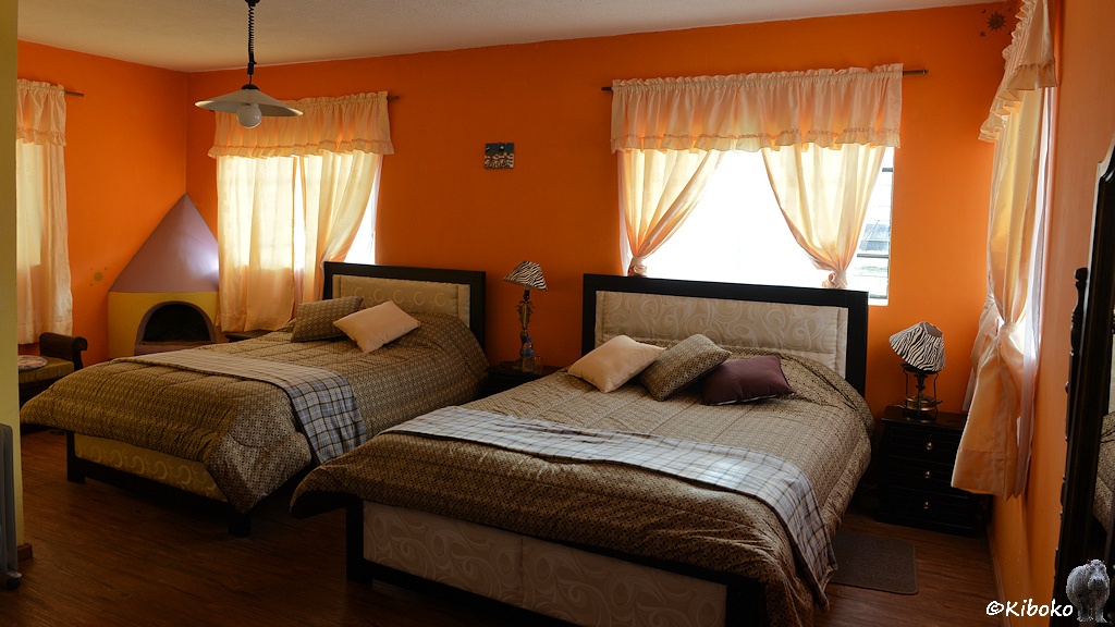 Das Bild zeigt die ein Zimmer in der Unterkaunft. Die Wände sind orange. Vor den vier Fenstern sind lichtdurchlässige orange Vorhänge. Im Zimmer stehen ein Doppelbett und ein Tripelbett. In der linken hinteren Ecke steht ein Kamin ohne Brennholz.