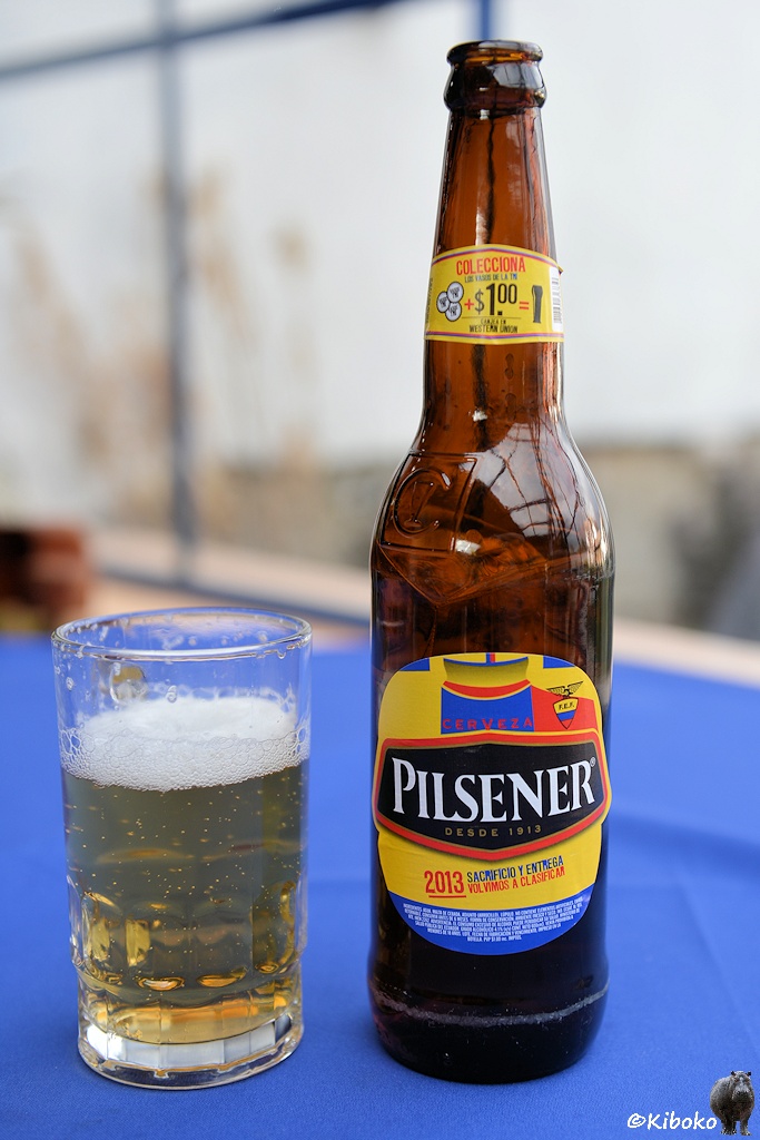 Das Bild zeigt eine Bierflasche mit einem Etikett in gelb-rot-blau mit der Aufschrift Pilsener. Daneben steht ein volles Bierglas.