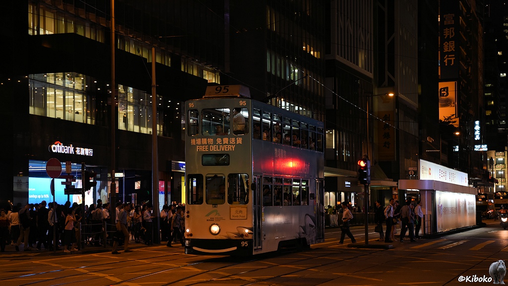 Das Bild zeigt eine Nachtaufnahme der weiß-hellblauen Tram 95 mit der Aufschrift: Free Delivery. Die Tram verläßt eine Haltestelle. Die Fußwege sind dicht bevölkert.