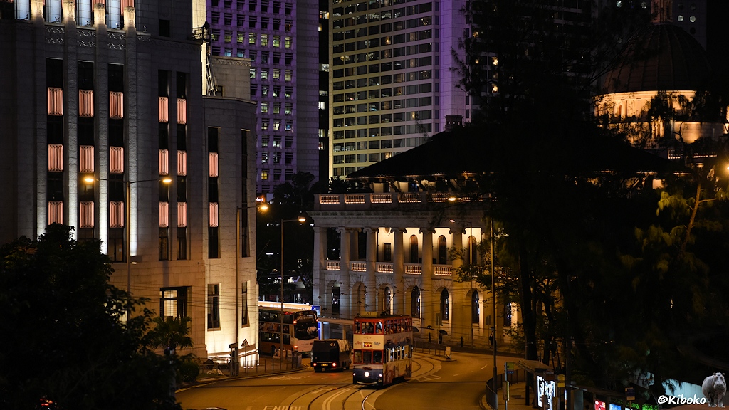 Das Bild zeigt eine Nachtaufnahme der rot-weiß-hellblauen Tram 51 in einer S-Kurve. Im Hintergrund ist ein älteres, monumentales Sandsteingebäude. Im Hintergrund sind lila angestrahlten Fassaden von Hochhäusern.