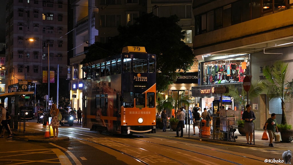 Das Bild zeigt die dunkelblau-ornage Tram 79 mit der Aufschrift Jupiter an einer beleuchteten Haltestelle in einer Geschäftsstraße. Am rechten Bidlrand ist ein Klamottenladen mit zweigeschossigen Schaufenstern.