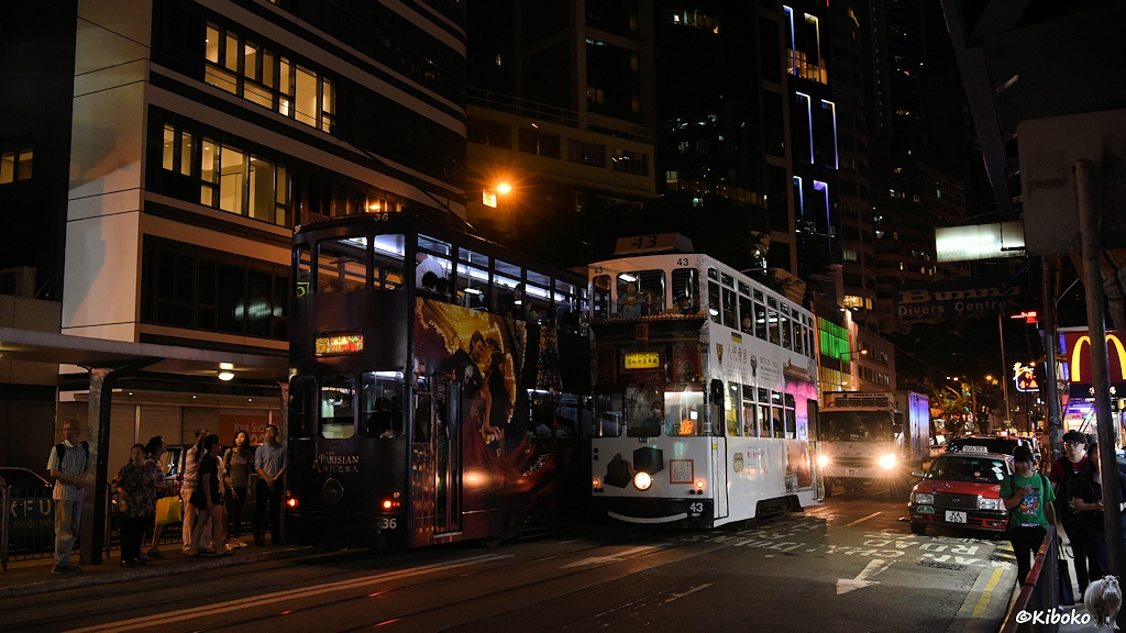 Das Bild zeigt die Begegnung der weißen Tram 43 mit der dunklen Tram 36 an einer Haltestelle in einer nächtlich beleuchteten Geschäftsstraße.