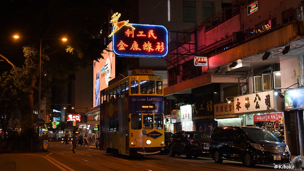 Das Bild zeigt die weiße Tram mit dunkelblauer Bauchbinde in einer Geschäftsstraße. Über der Tram ist ein große Leuchtreklame mit chinesischen Schriftzeichen und einen springenden Hirsch.