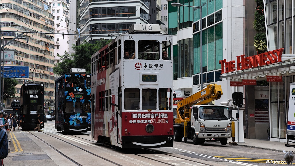 Das Bild zeigt weiß-rote doppelstöckige Straßenbahn mit der Nr. 113 auf einer vierspurigen Straße. Es folgt eine schwarze 104 Straßenbahn mit blauen Blumen muster. Die schwarze Tram 82 verschwindet auf dem Nachbargleis.