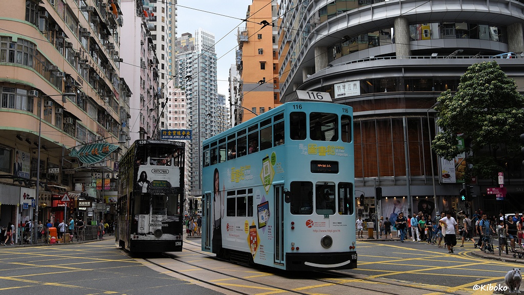 Das Bild zeigt eine hellblaue Tram mit der Nummer 116 beim überqueren einer Kreuzung. Rechts daneben fährt die schwarze Tram 138 in die Gegenrichtung. Auf der Kreuzung ist ein Rautenmuster aus gelben Streifen. Neben der Kreuzung sind hohe Geschäftshäuser. Im Hintergrund stehen Hochhäuser.