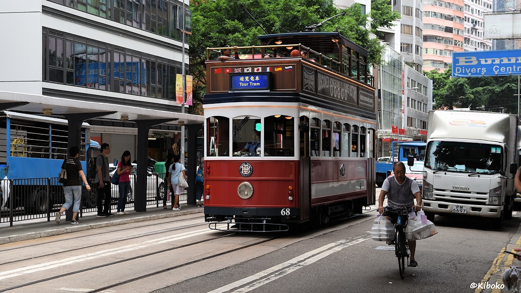 Das Bild zeigt eine weiß-rote Tram mit der Nummer 68, die ein offenes Oberdeck hat. Die Tram für Touristen fährt an einer Haltestelle vorbei. Im Vordergrund ist ein radfahrender Essenbringdienst mit vielen Plastiktüten am Lenker.