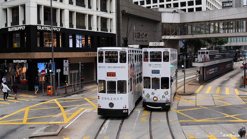 Das Bild zeigt die Begegnung von zwei weißen Trams mit den Nummern 152 und 142 auf einer Kreuzung. Im Hintergrund ist eine Haltestelle, Geschäftshäuser und eine Verbindungsbrücke zwischen den Straßenseiten.