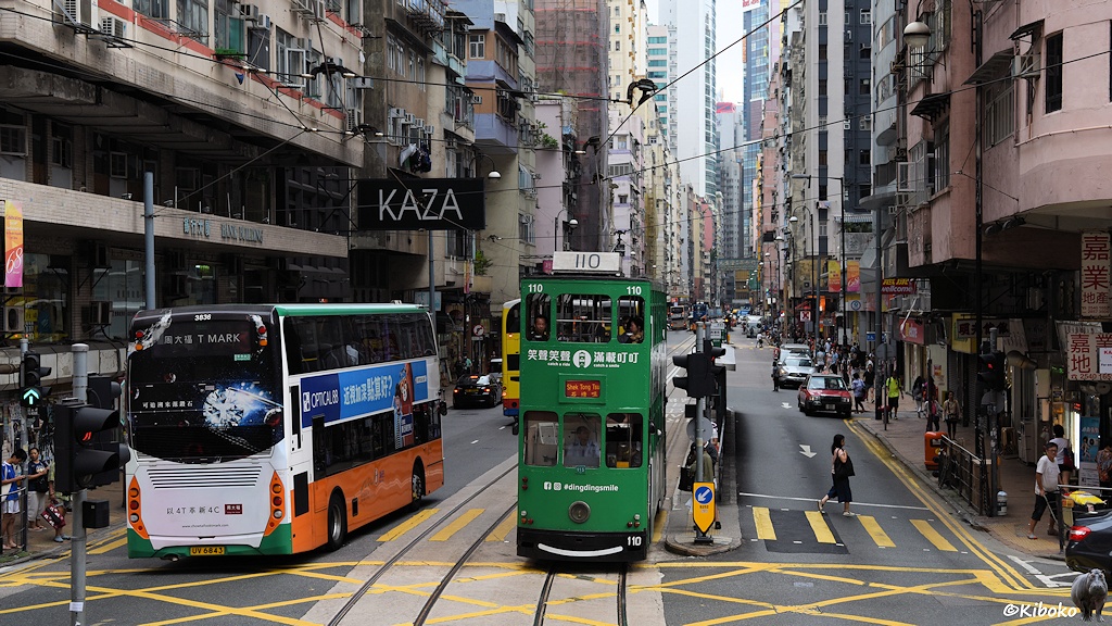 Das Bild zeigt  die grüne Tram 110 zwischen doppelstöckigen Bussen auf einer Geschäftsstraße an einer Kreuzung.