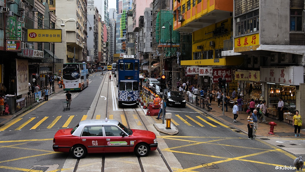 Das Bild zeigt die blaue Tram 133 an einer Haltestelle in einer Geschäftsstraße. Im Vordergrund kreuzt ein rot-weißes Taxi die Gleise.