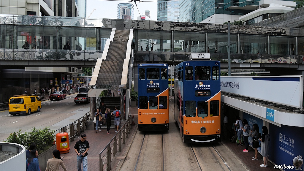 Das Bild zeigt zwei begegnende durnkelblau-orange Trams mit den Nummern 42 und 79 an einer Haltestelle. Die Zugänge der Haltestelle sind von einer überdachten Brücke mit langen Treppen.