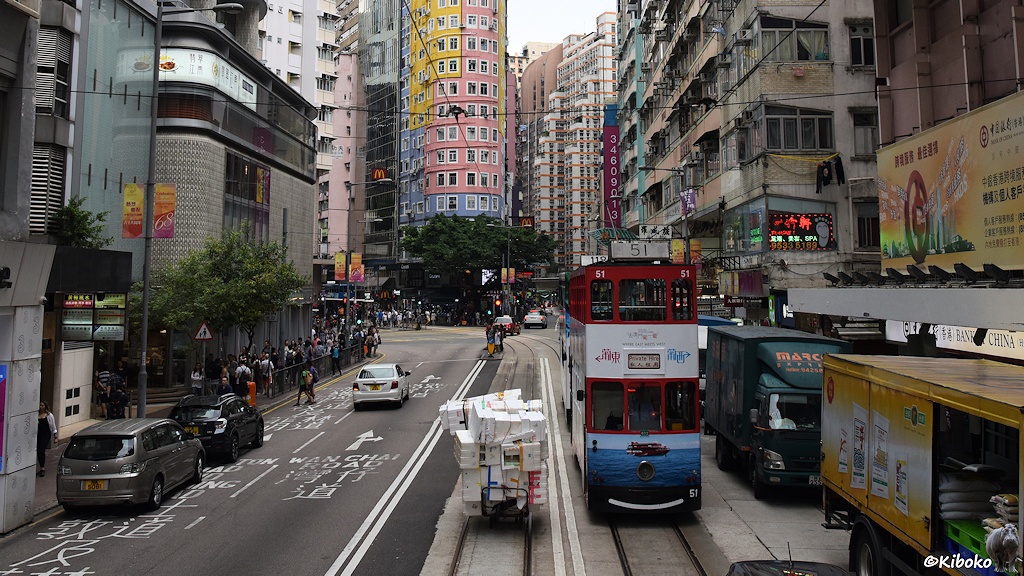 Das Bild zeigt die rot-weiß-blaue tram 51 auf einer Geschäftsstrße. Links blockieren LKWs beim Entladen die Fahrbahn. Vor der tram zieht ein Mann einen Handwagen mit einen großen Berg mit Styropor.