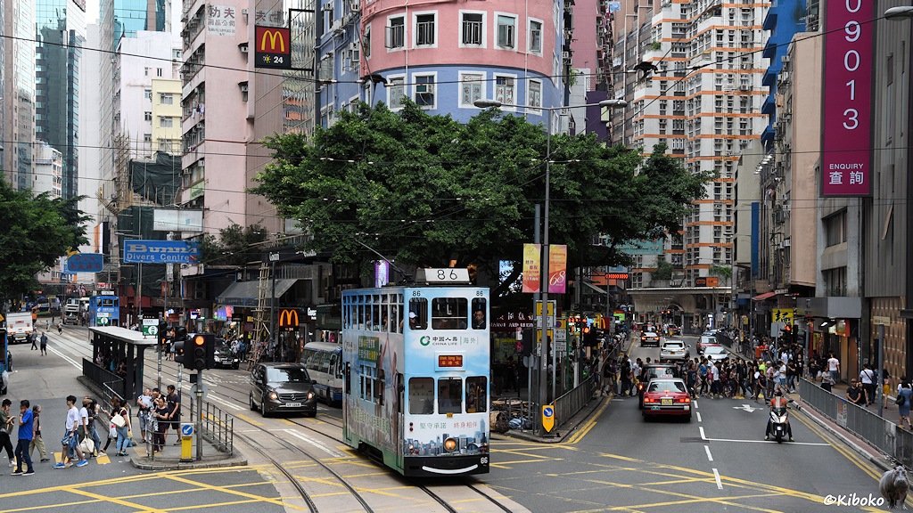 Das Bild zeigt die hellblau-weiße Tram 86 beim überqueren einer Kreuzung in einem Geschäftsviertel.