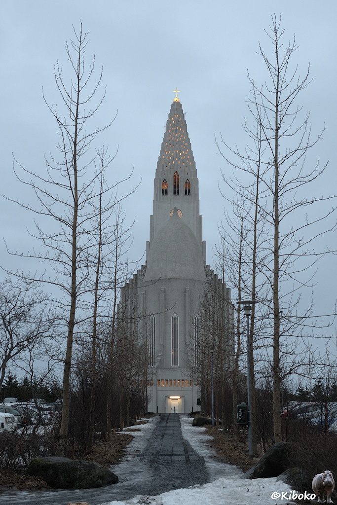 Hochkantaufnahme einer hellgrauen Kirche durch eine Allee laubloser Bäume.