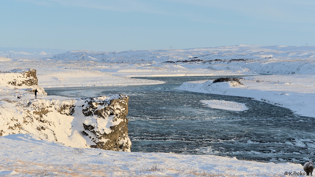 Das Bild zeigt einen blauen Fluss mit hellen Kronen, der in S-Kurven durch ein schneebedecktes Tal fließt. Auf der linken Seite sind an einer Felsformation zwei Fotografen.