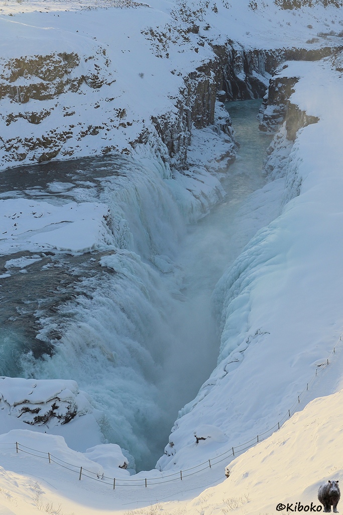 Das Bild zeigt eine Detailfaufnahme der unteren Stufe des Wasserfalls und der Schlucht im Hochformat. Hellblaues Eis säumt beide Seiten der Schlucht. Unterhalb des Wasserfalls ist das abfließende Wasser durch die Gisch nicht zu sehen. 