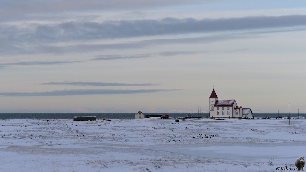 Das Bild zeigt eine kleine weiße Kirche mit rotem Dach auf einer weiten, verschneiten Ebene. Dahinter ist im dunklen Türkis das Meer.
