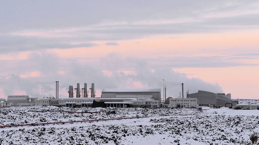 Das Bild zeigt mehrere Industriegebäude auf einer verschneiten Felsebene. Aus einem Kühlturm steigt Dampf auf, den der Wind über die Anlage weht.