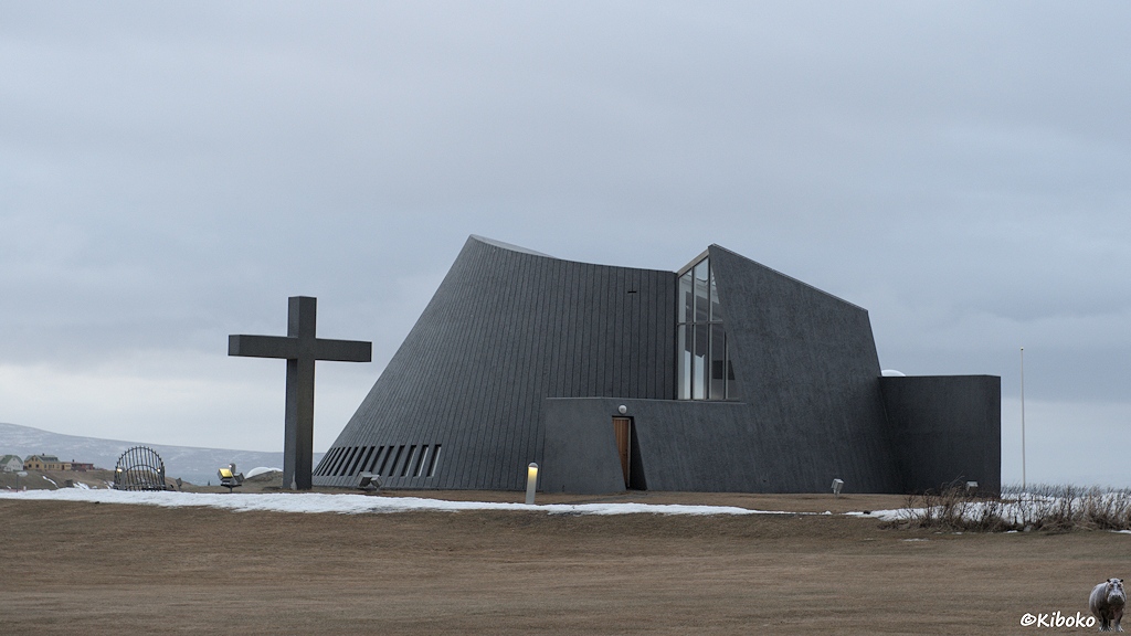 Ein schwarzes Gebäude mit schrägen und geschwungenen Wänden. Davor steht ein überdimensionales Kreuz.