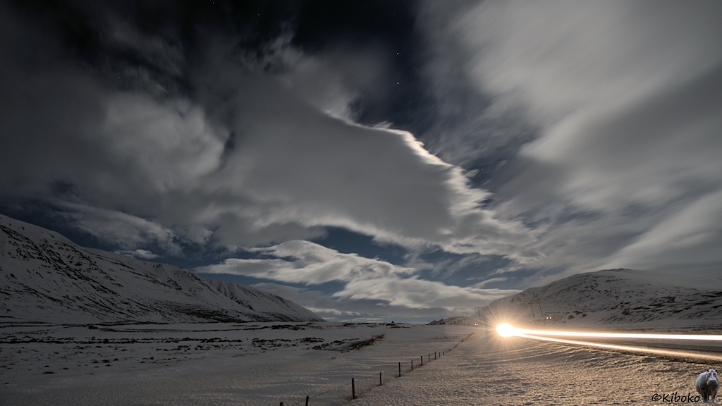 Der Wind jagt Wolken über den Nachthimmel. Dazwischen leuchten Sterne über ein schneebedecktes Tal. Am rechten Bildrand ist eine Straße, die durch Autoscheinwerfer beleuchtet wird.
