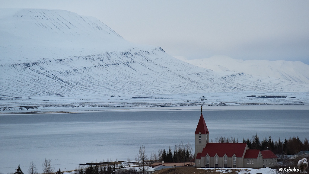 Eine Kirche aus Beton mit spitzen rotem Dach überragt ein Gewässer. Am gegenüberliegenden Ufer steigen schneebedeckte Berge empor.