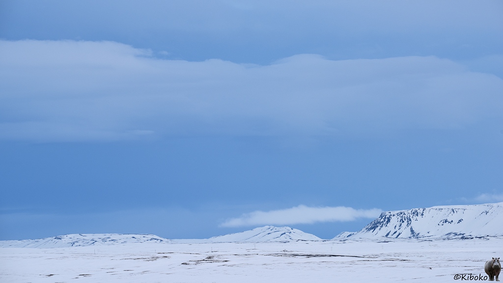 Blaugraue Wolken hängen über einer verschneiten baumlosen Ebene. Ein graues und ein weißes Wolkenband ziehen sich über den Himmel. Am Horizont sind ein paar Berge.