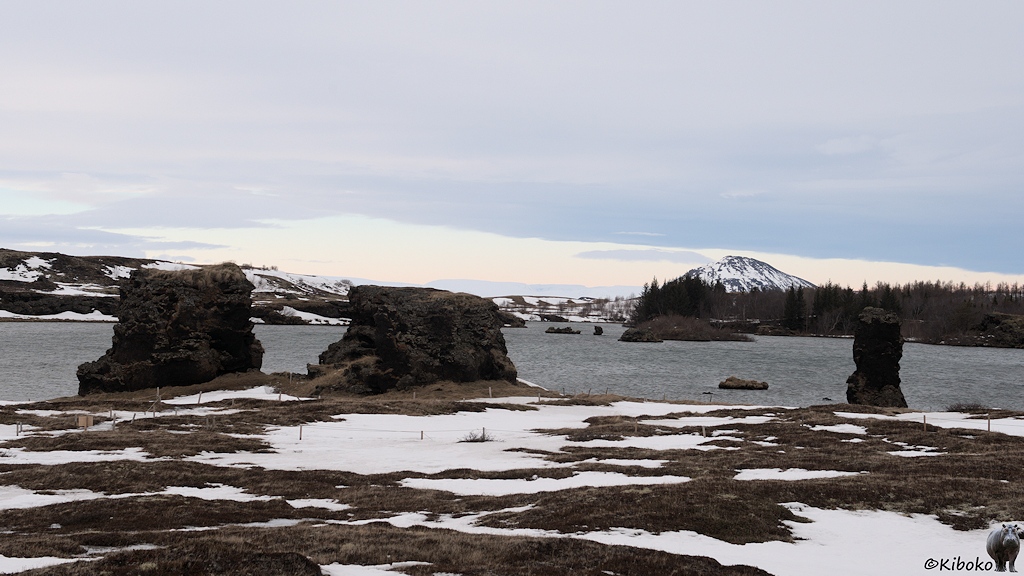 Drei dunkle Steinskulpturen stehen an einem See. Im See sind zahlreiche kleine Felsinseln. Auf einer Halbinsel im Hintergrund stehen Bäume.