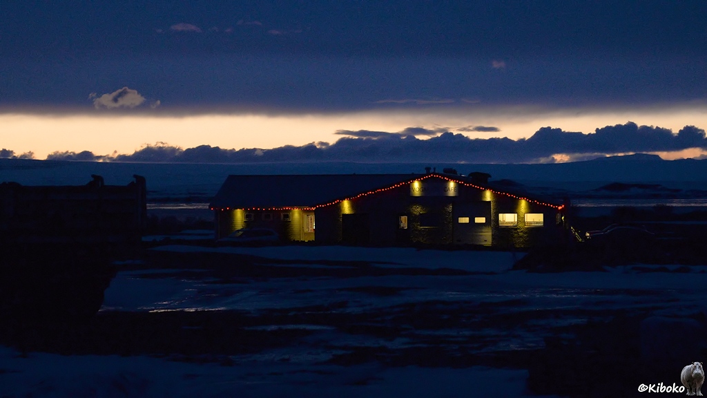 Eine braune Holzhütte mit roter Lichterkette am Giebel, steht an einem See zur blauen Stunde.