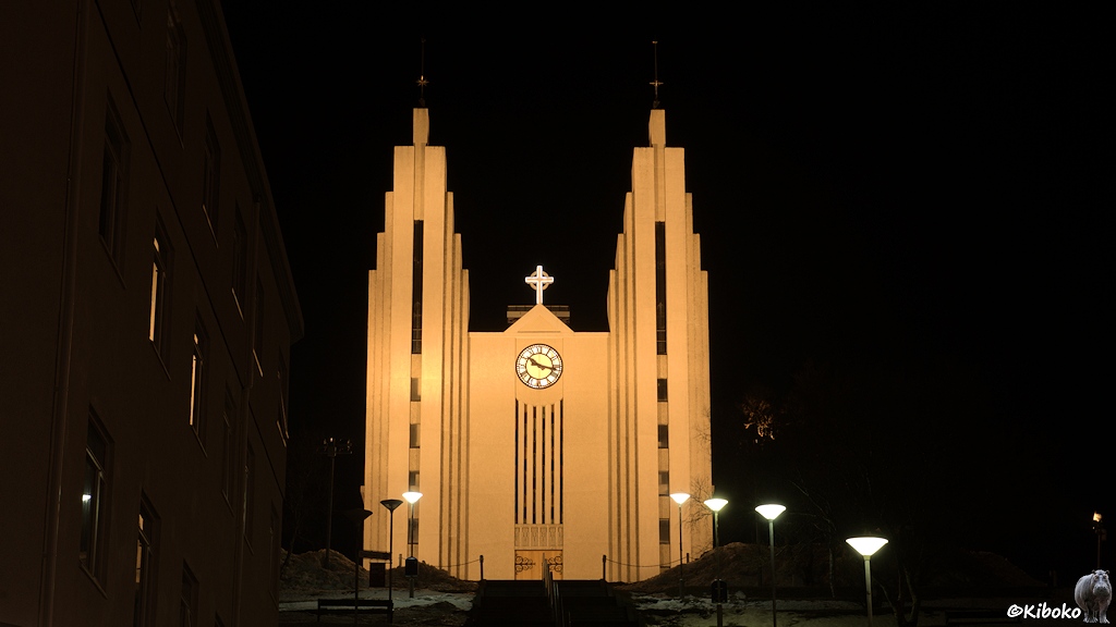 Eine Kirche mit zwei Betontürmen aus eckigen Säulen wird nachts angestrahlt. Eine beleuchtete Treppe führt aufwärts zum Kircheneingang.