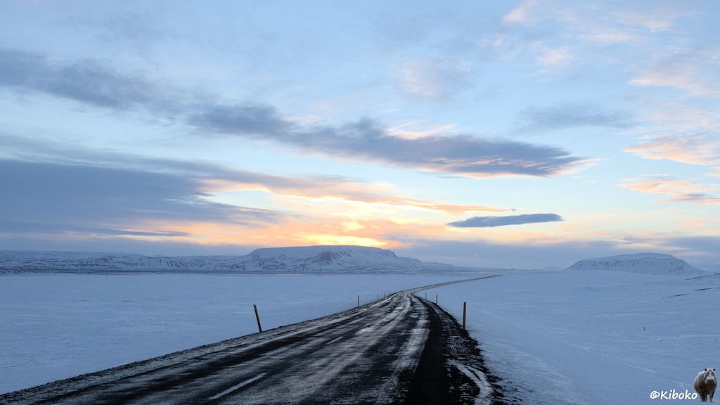 Eine schwarze Teerstraße führt über eine weiße, schneebedeckte Ebene. Hinter den schneebedeckten Bergen im Hintergrund geht die Sonne auf.
