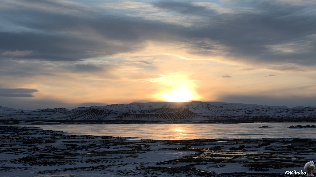 Die Sonne geht über einer Bergkette und einen Vulkankrater auf. Darunter befindet sich ein zugefrorener See. Sie Sonne spiegelt sich im Eis.