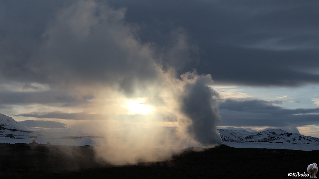 Eine Fumarole mit aufsteigenden Dampf im Gegenlicht. Die Sonne steht gerade so über einer verschneiten Bergkette im Hintergrund.