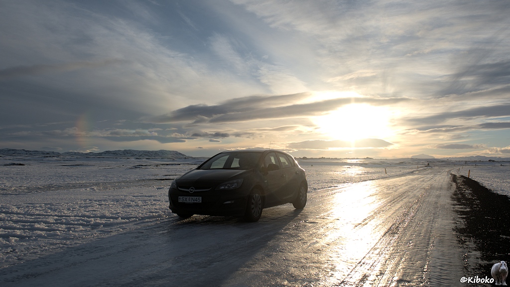 Ein dunkles Auto steht auf einer vereisten Straße auf einer verschneiten Ebene. Die Sonne spiegelt sich auf dem Eis auf der Straße.
