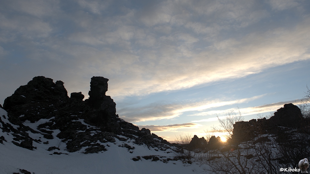 Dunkle Säulen aus Lava stehen im Gegenlicht der untergehenden Sonne. Dazwischen sind Schneefelder und kahle Büsche.
