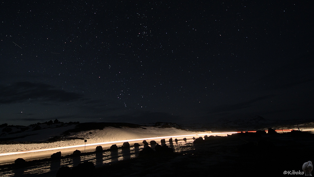 Nachtaufnahme einer Straße, die durch durchfahrende Autos beleuchtet wird. Darüber ist ein Nachthimmel mit vielen Sternen. Die Landschaftsstruktur mit Lavsäulen zeichnet sich schwach gegen den Nachthimmel ab.