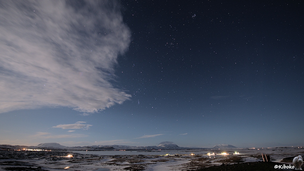 Nachtaufnahme eines gefrorenen Sees in einer weiten Winterlandschaft. Ein Wolkenband schiebt sich vom linken Bildrand ins Bild.