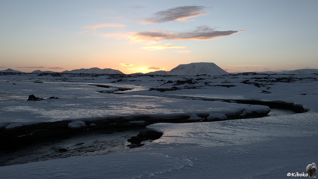 Ein mäandrierender Warmwasserfluss führt über eine verschneite Hochebene. Im Hintergrund ist eine Bergkette. Hinter der Bergkette ist ein heller Punkt, der den Sonnenaufgang ankündigt.