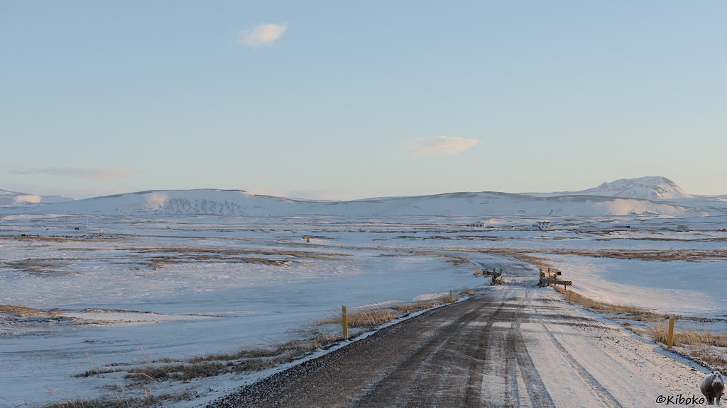 Eine schmale, schneebedeckte Straße führt über eine kleine Brücke durch eine einsame Winterlandschaft.