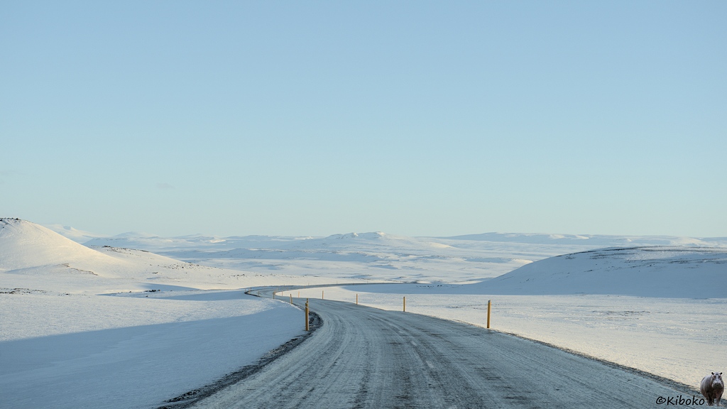Eine vereiste Straße führt mit einem S-Bogen durch ein schneebedecktes Tal. An den Seiten und im Hintergrund sind schneebedeckte Berge