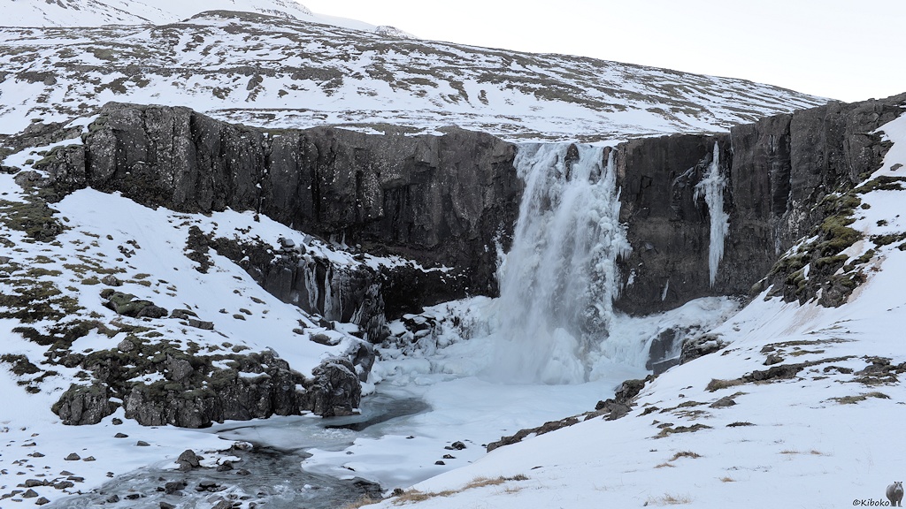 Ein Wasserfall stürzt sich über eine halbrunde, rund 15m hohe Felskannte. Neben dem Wasserfall sind hängen lange Eiszapfen. Der Fluss ist vereist.