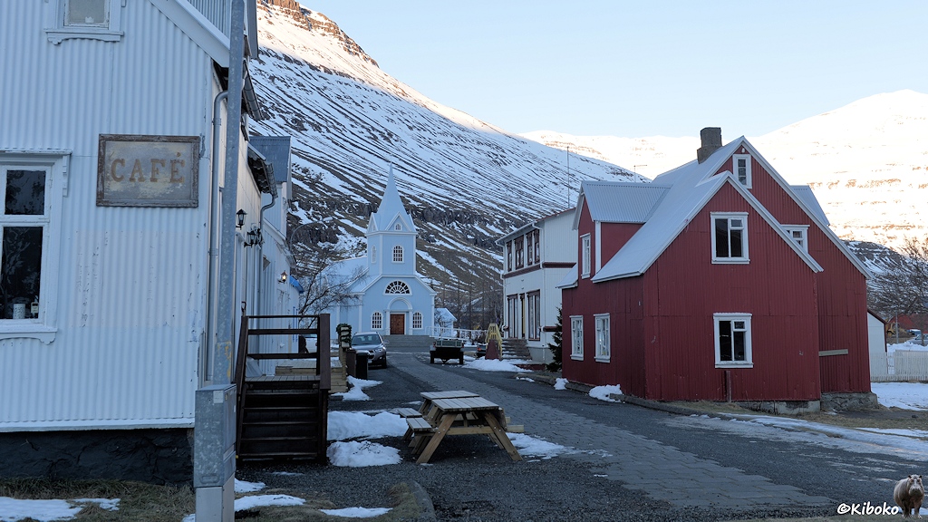 Weiße und rotbraune Holzhäuser säumen einen Weg. Am Ende steht eine hellblaue Kirche.