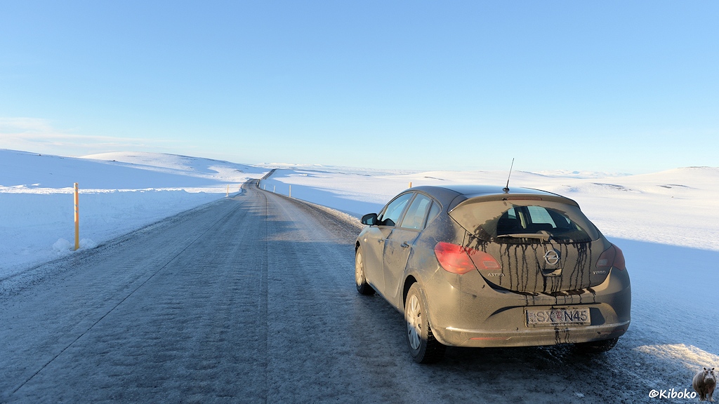 Ein schwarzes, total verdrecktes Auto steht auf einer vereisten Straße in einer verschneiten Bergwelt.