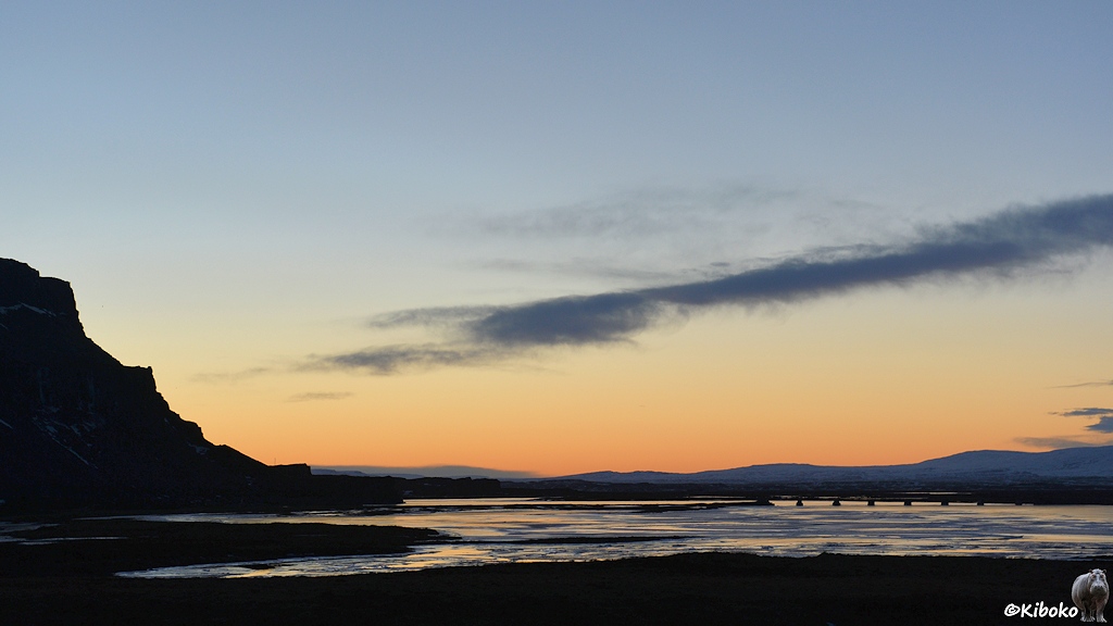 Eine Schwemmlandschaft im spätabendlichen Gegenlicht. Der Horizont schimmert orange. Eine Brücke führt über einen gefrorenen Fluss. Die Silhouette eines Berges ist am linken Bildrand.