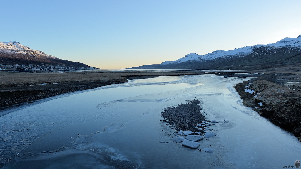 Ein teilweise zugefrorener Fluss fließt vom Betrachter weg in einem Fjord. Der Fluss und der Fjord liegen im Schatten.