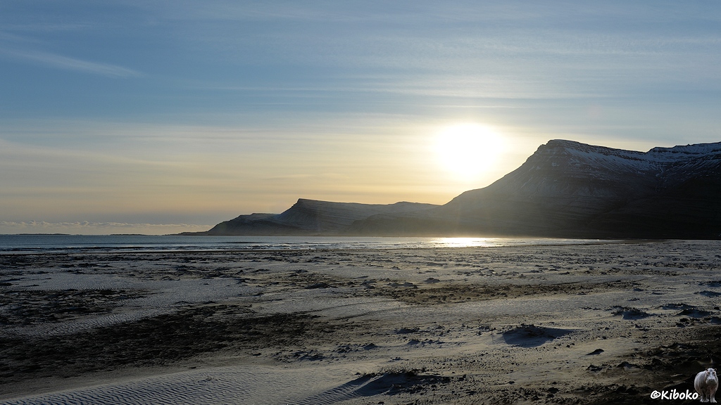 Sandstrand an einer breiten Bucht im Gegenlicht. Die tiefstehende Mittagssonne strahlt über eine Bergkette.