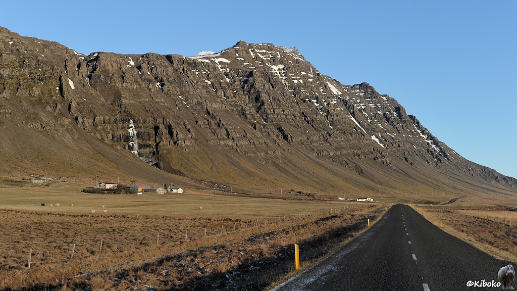 Eine Teerstraße führt ins Bild. Auf der linken Seite sind am Fuße einer Bergkette kleine Bauernhöfe. Ein Wasserfall verziert die Bergkette. Auf den eingezäunten Wiesen weiden Schafe.