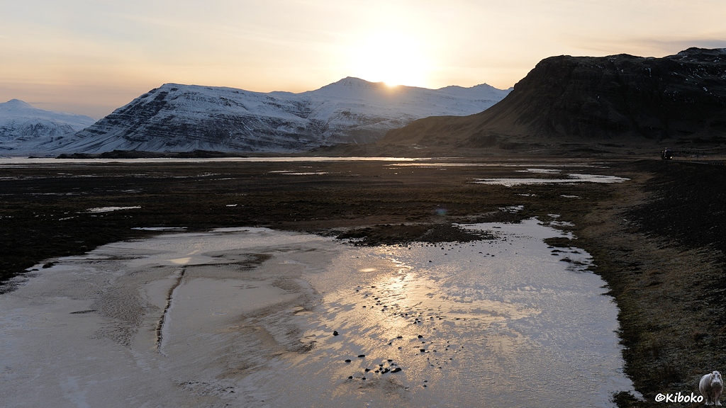 Das Bild zeigt eine schwarze Schotterebene mit weißen zugefrorenen Pfützen. Auf einer Eisfläche im Vordergrund spiegelt sich die Sonne, die im Hintergrund gerade hinter einer schneebedeckten Bergkette verschwindet.
