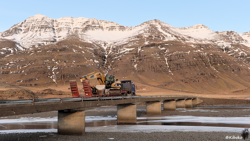 Das Bild zeigt eine lange Betonbrücke mit 6 Pfeilern. Auf der Brücke fährt gerade ein LKW mit einem Anhänger auf dem ein Bagger geladen ist. Die Brücke führt über einen flachen und breiten Flusslauf. Im Hintergrund sind sonnenbeschienene Berge, die oben mit Schnee bedeckt sind.
