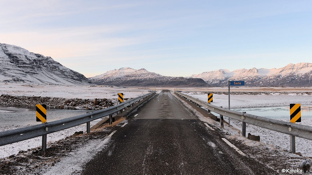 Das Bild zeigt eine Straße über eine einspurige Brücke. Auf beiden Seiten sind Leitplanken und gelb-schwarze Warnschilder. Die Brücke führt über einen Fluss. Im Hintergrund scheint die Sonne auf schneebedeckte Berge.